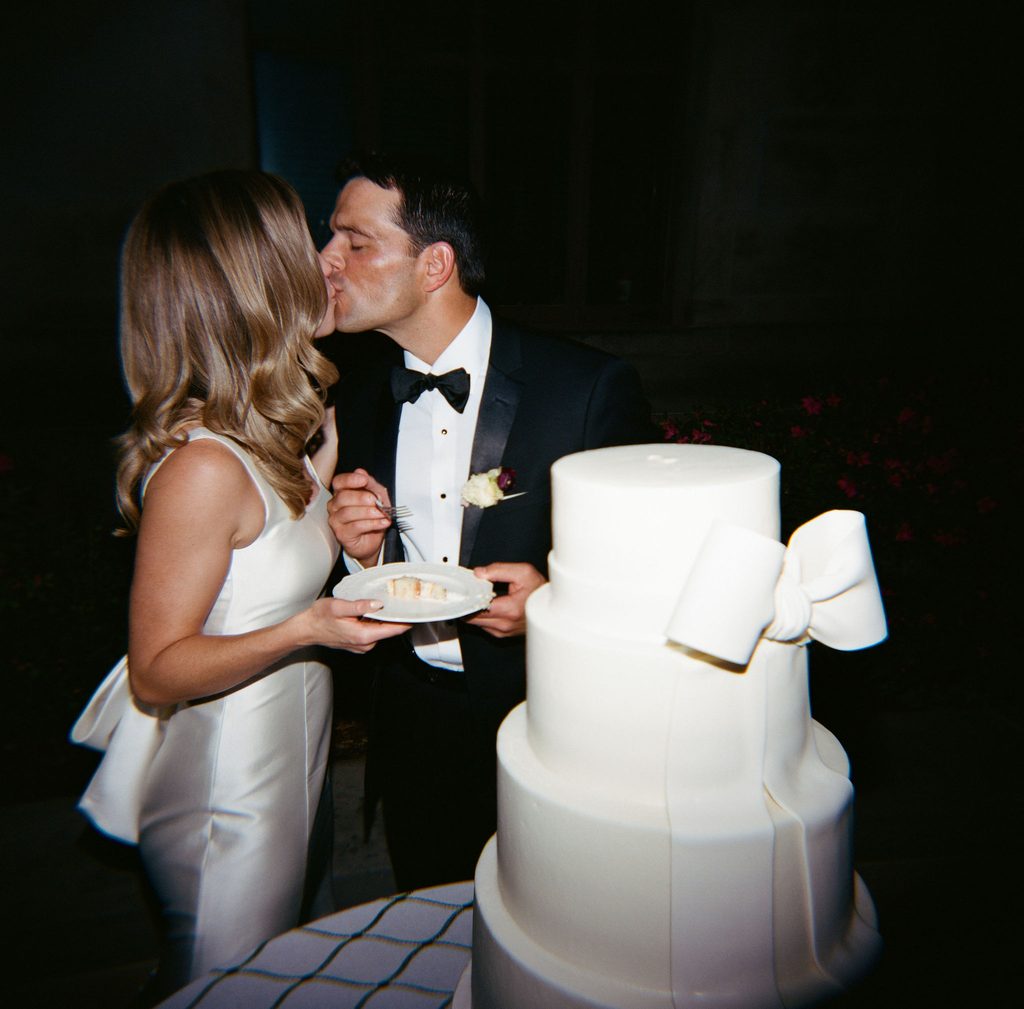Bride and groom enjoying a slice of multi-tiered white wedding cake with bow detail by Amy Cakes at Oklahoma Hall of Fame reception.