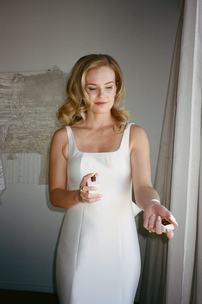 Bride in square-neck wedding gown applying perfume in bridal suite at Gaylord-Pickens Museum venue.