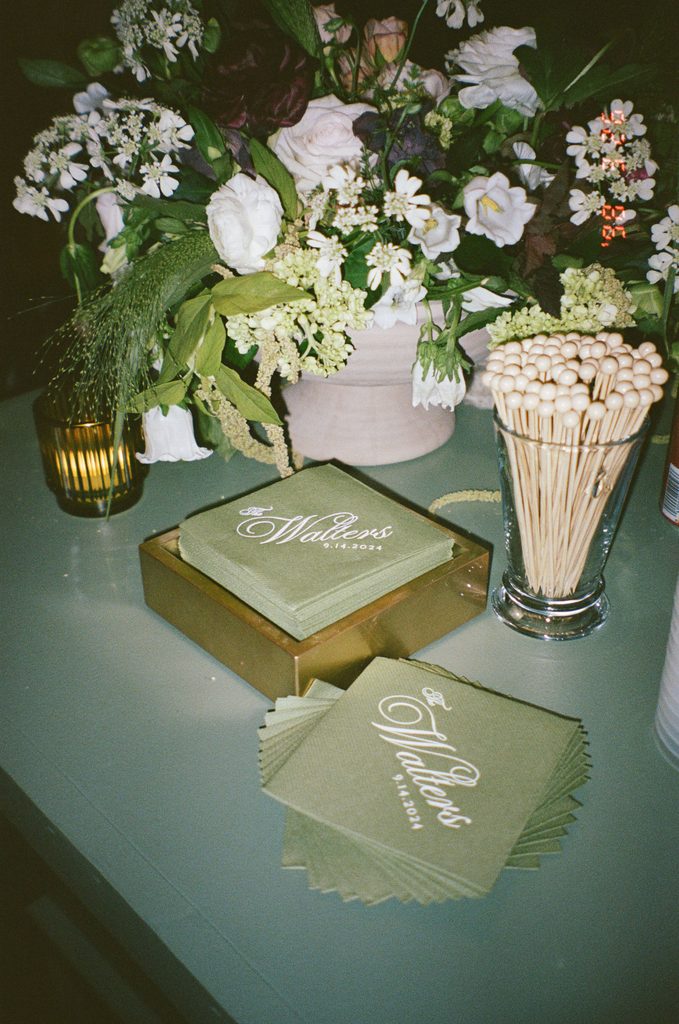 Close-up detail of wedding guest book and green linen napkins on reception table with white floral arrangement by Emmaline Rose Floral.