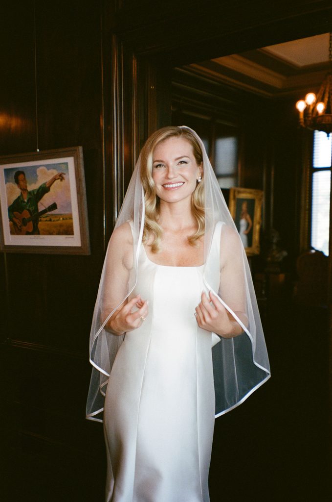 Bride posing in indoor doorway with cathedral veil framing square-neck satin gown at Gaylord-Pickens Museum venue.