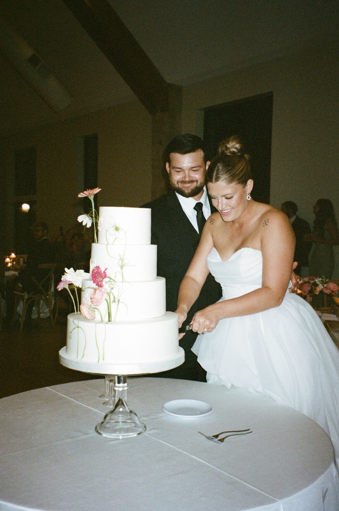 Bride cutting a multi-tiered white wedding cake adorned with fresh flowers at The Bower venue reception in Edmond, Oklahoma.