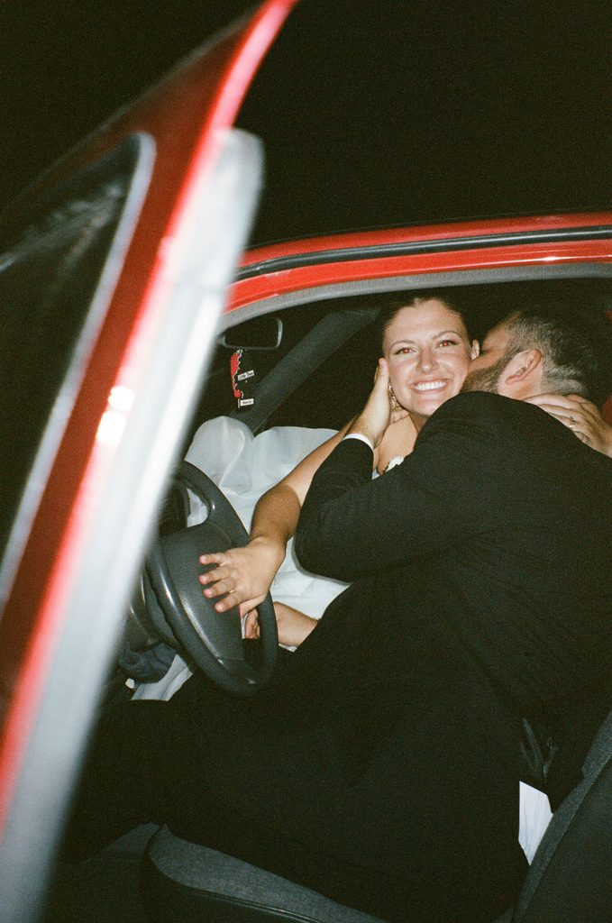 Candid flash photograph of groom kissing bride inside a red vintage get-away car after wedding reception at The Bower.
