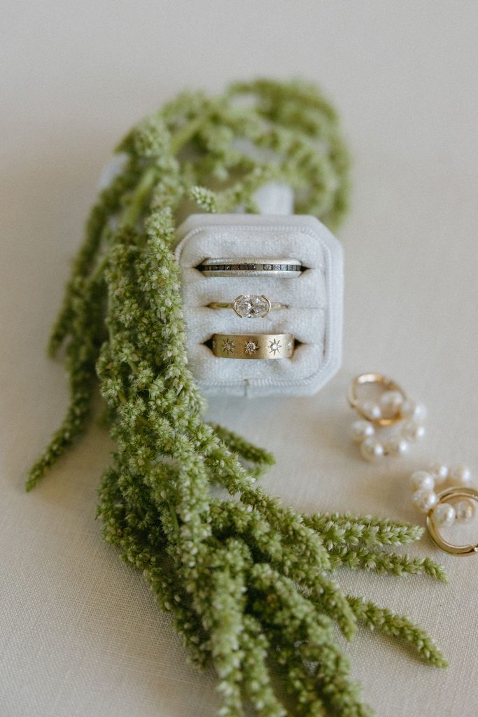 Close-up photo of oval engagement ring and gold wedding bands inside a velvet ring box with greenery detail.