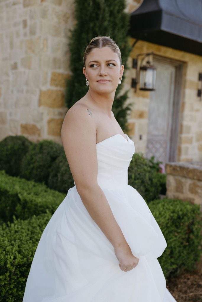 Close-up photo of bride holding up layered white wedding dress skirt with green manicured hedge in background.