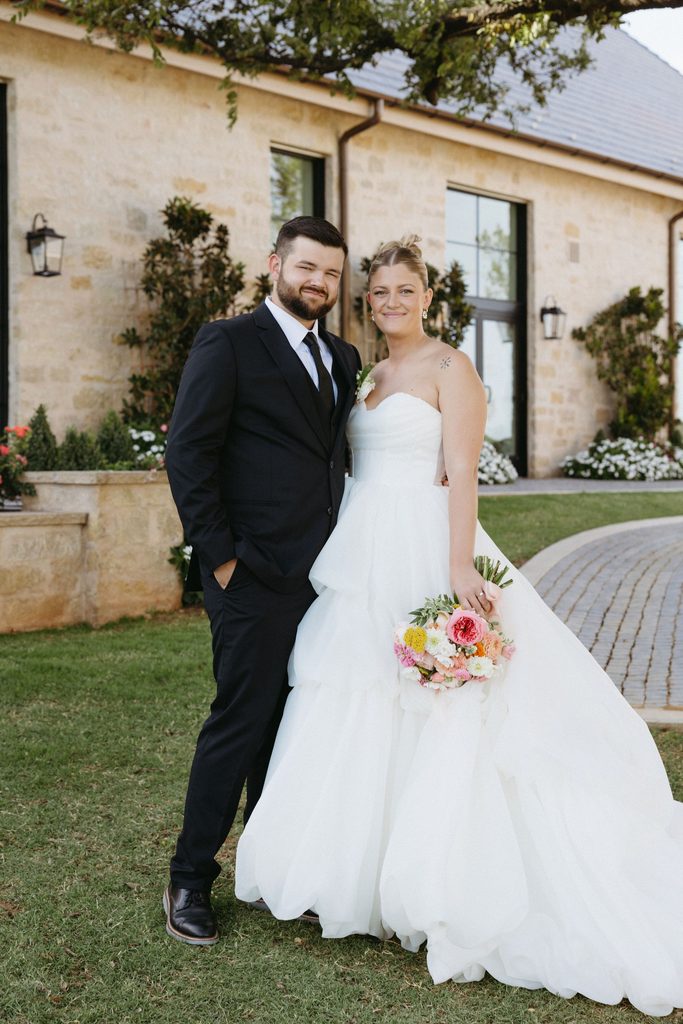 Full-length portrait of bride in strapless layered tulle wedding dress and groom in black suit standing together near limestone courtyard at The Bower in Edmond, Oklahoma.