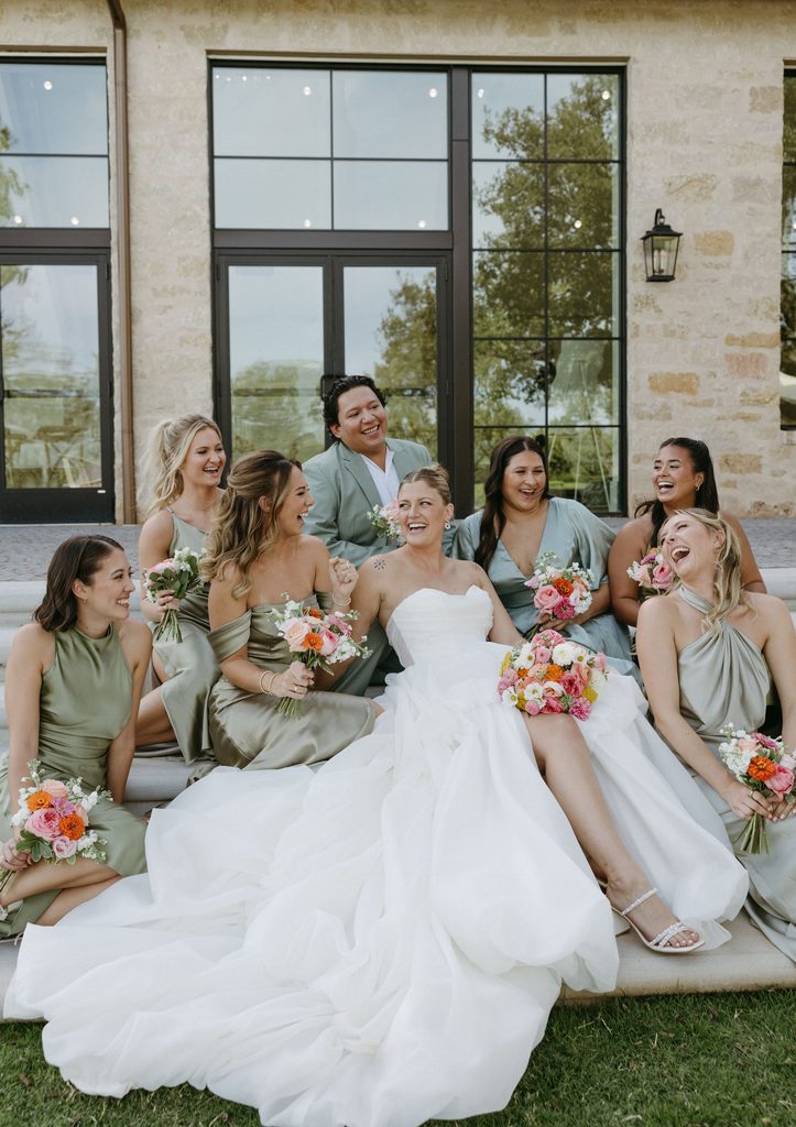 Bride in strapless ballgown laughing with bridesmaids in sage green dresses holding colorful coral and yellow floral bouquets outdoors.