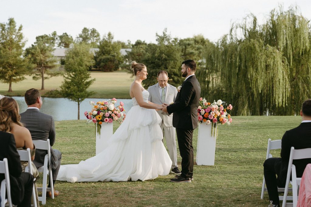 Bride and groom exchanging vows during an outdoor waterfront wedding ceremony officiated at The Bower with colorful floral pillars.