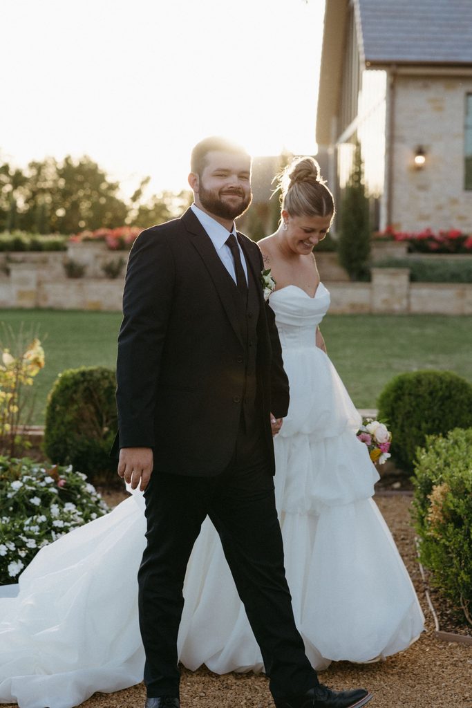 Full-length portrait of bride and groom walking hand-in-hand through garden grounds at The Bower wedding venue in Edmond, OK.