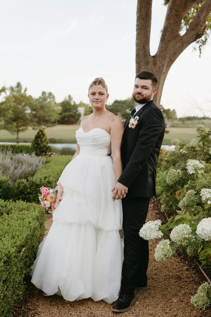 Bride and groom standing together under large trees outdoors at waterfront wedding venue, The Bower in Edmond, OK.
