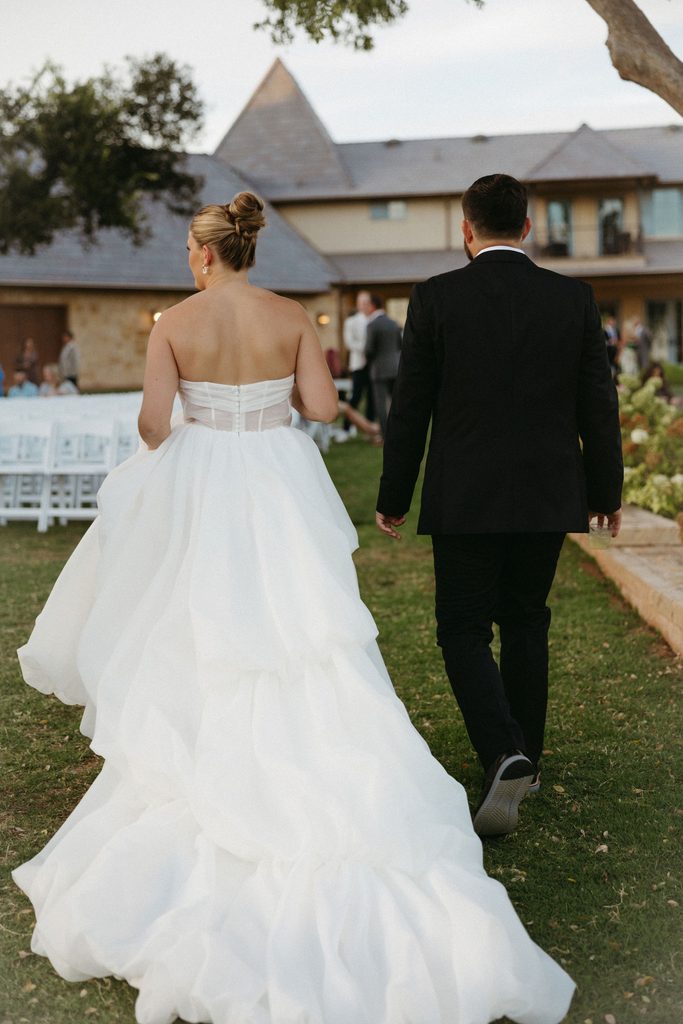 Bride and groom walking hand-in-hand across lawn toward limestone pavilion during outdoor wedding reception at The Bower in Edmond, OK.