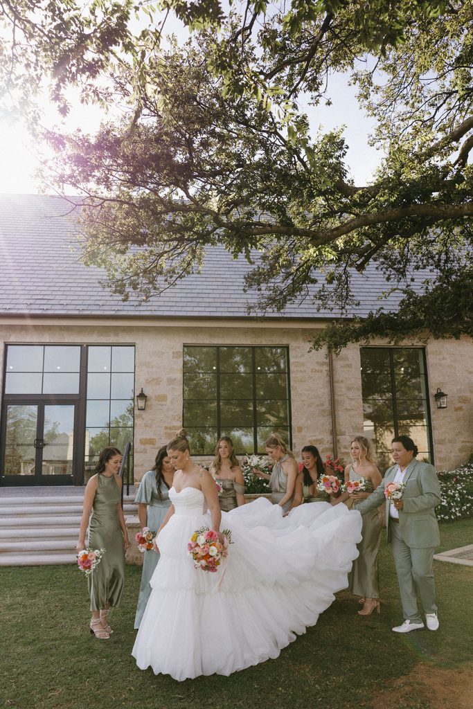 Bride holding up layered tulle wedding dress with bridesmaids in sage dresses outside limestone building at The Bower in Edmond, Oklahoma.