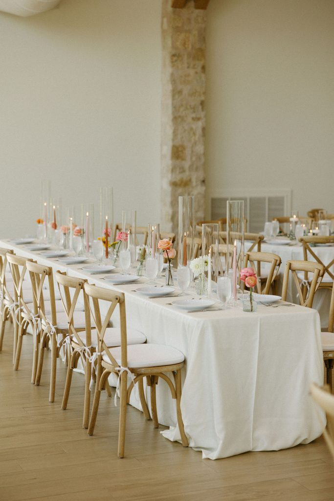 Indoor wedding reception long head table set with white linens, cross-back chairs, and tall clear flower vases at The Bower venue.