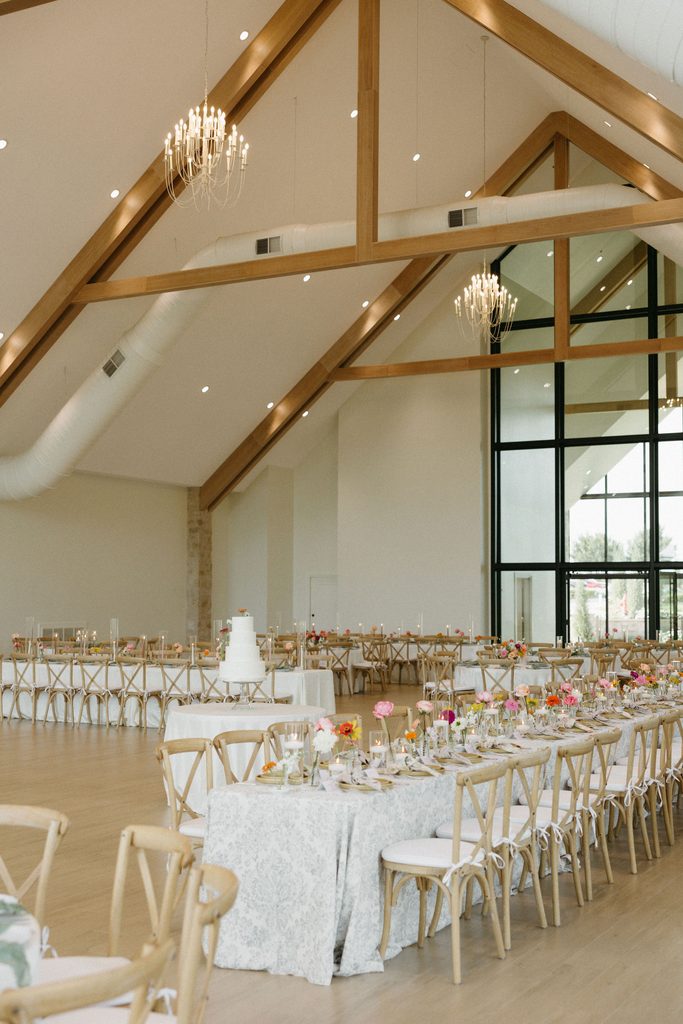 Indoor wedding reception setup at The Bower featuring wooden beam ceilings, long tables with white linens, cross-back chairs, and bright floral centerpiece bud vases.