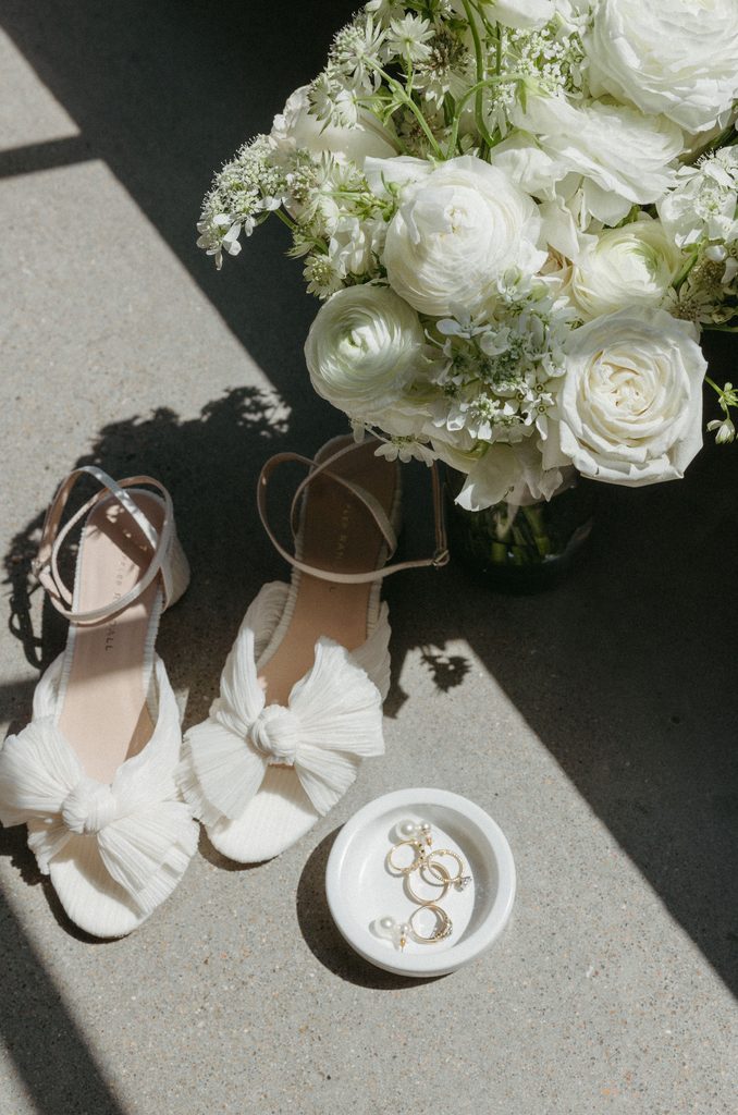 Bride's satin heels with oversized bows sitting in sunlight next to ring dish and white rose floral arrangement at Oklahoma Hall of Fame.