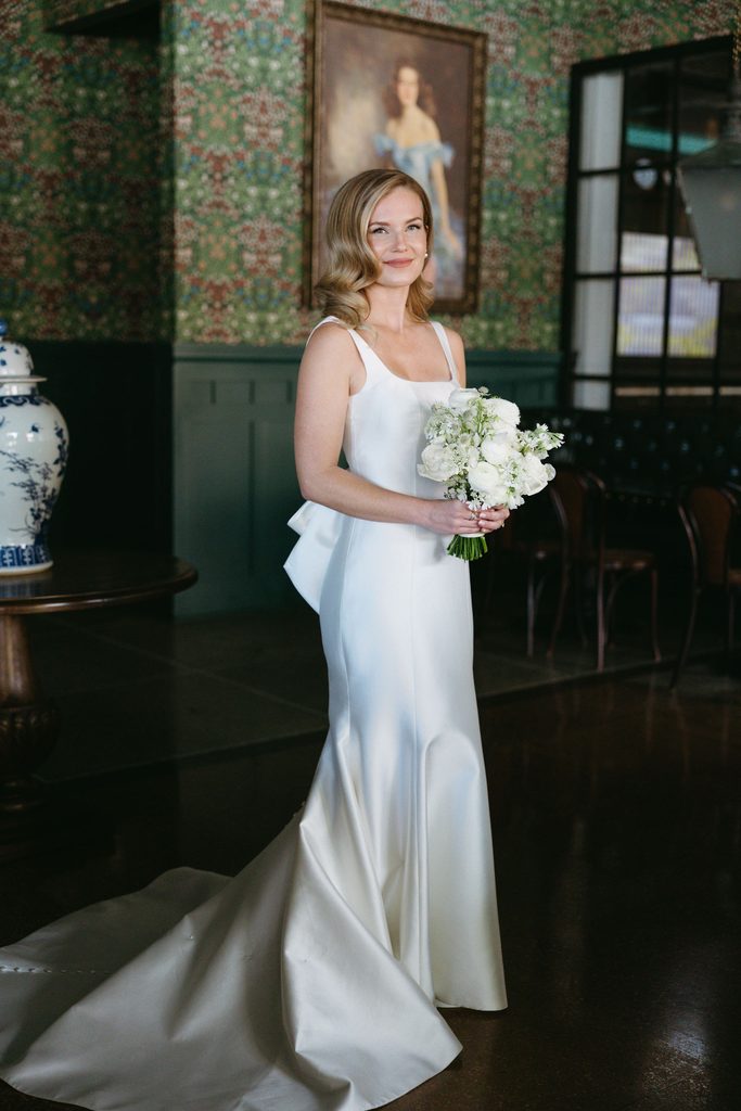 Bride in square-neck satin Neiman Marcus wedding gown posing in green wallpaper bridal suite at Gaylord-Pickens Museum.
