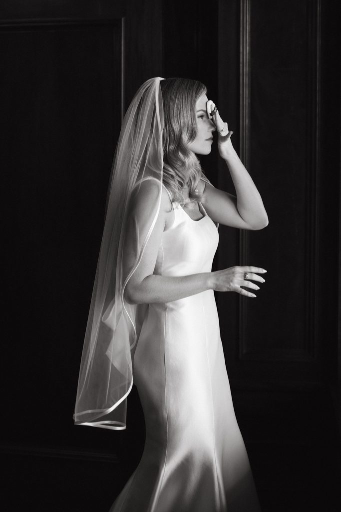 Black and white portrait of bride adjusting veil framing square-neck Neiman Marcus dress at Oklahoma Hall of Fame.