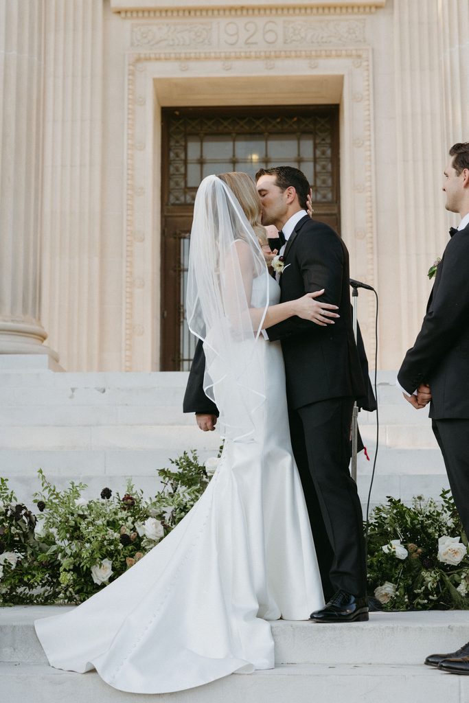 Bride and groom kissing on stone steps framed by columns during outdoor ceremony at Gaylord-Pickens Museum venue.