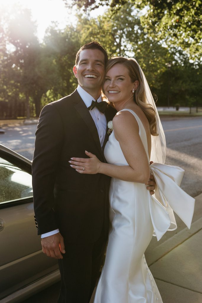 Smiling bride and groom in black tie attire posing outdoors together beside car at Gaylord-Pickens Museum in Oklahoma City.