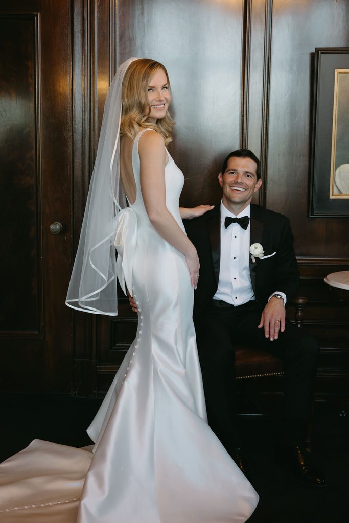 Bride in square-neck satin Neiman Marcus wedding gown with bow detail sitting with groom on indoor bench at Oklahoma Hall of Fame.