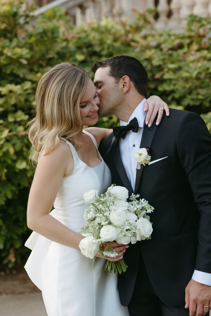 Groom in black bowtie kissing bride on the cheek outdoors at Gaylord-Pickens Museum venue in Oklahoma City.