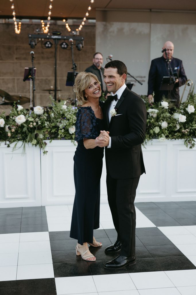Smiling bride and groom in black tie attire posing together on a black-and-white checkered dance floor during outdoor garden reception at Oklahoma Hall of Fame.