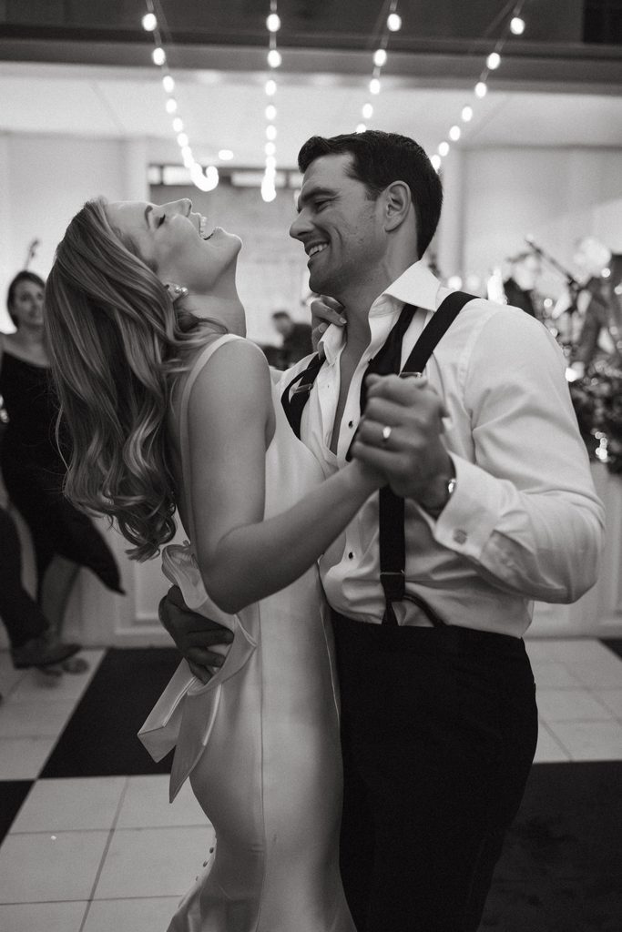 Candid black and white photograph of bride and groom laughing while dancing on checkered dance floor under string lights at Gaylord-Pickens Museum.