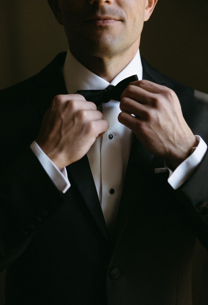 Close-up detail of groom adjusting black bowtie in black tie attire at Oklahoma Hall of Fame wedding.