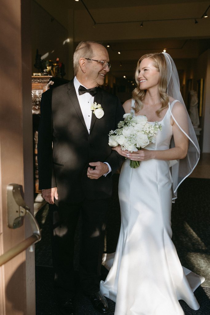 Bride in square-neck satin Neiman Marcus wedding gown smiling with her father in tuxedo before ceremony at Oklahoma Hall of Fame.