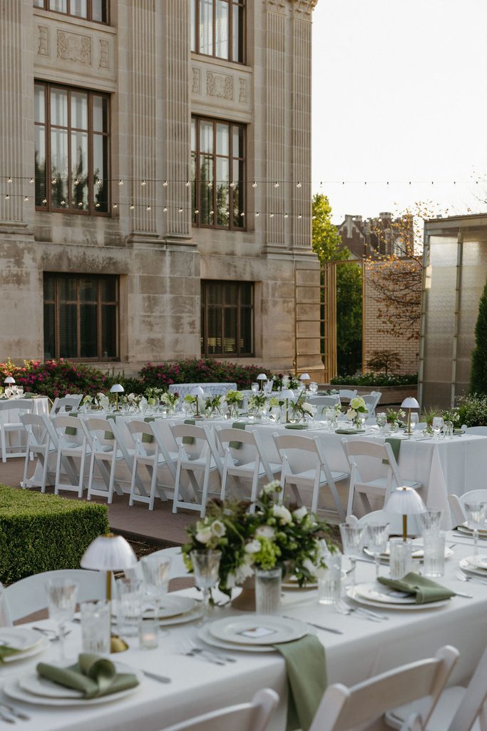 Long outdoor reception tables with white linens, green napkins, and white floral centerpieces set in garden courtyard at Oklahoma Hall of Fame.