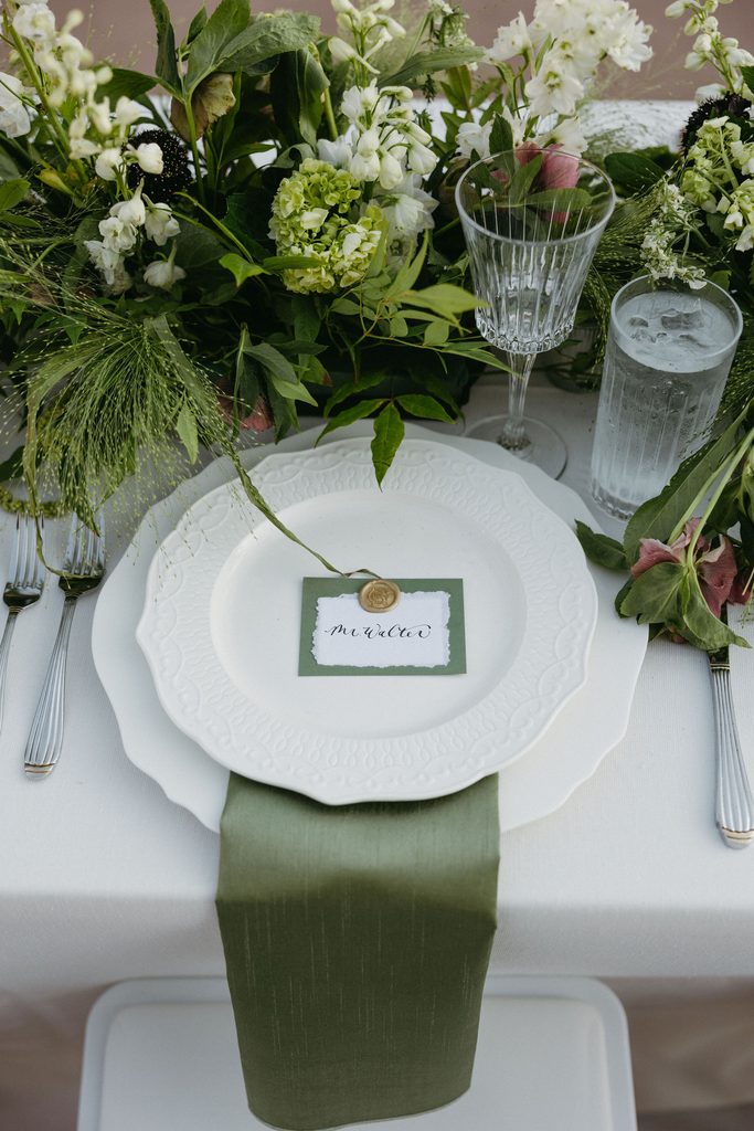 Wedding reception place setting with green linen napkin, place card, and white floral centerpieces by Emmaline Rose Floral.
