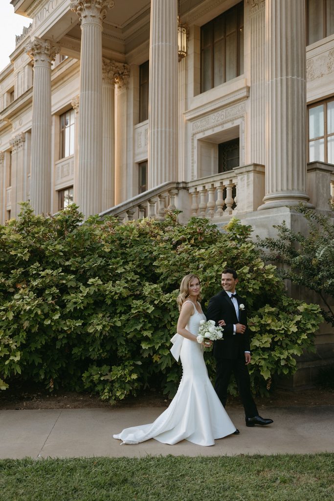 Bride in satin Neiman Marcus wedding dress with oversized back bow standing outside historic Oklahoma Hall of Fame building with grand stone columns.