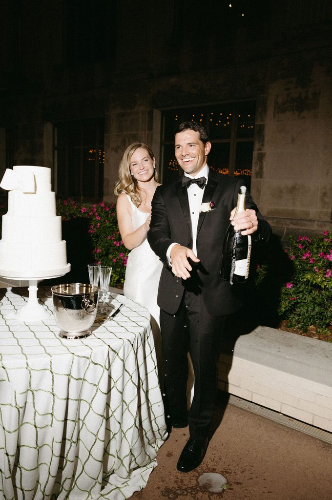 Bride and groom cutting multi-tiered white wedding cake by Amy Cakes with champagne bottle during evening reception at Oklahoma Hall of Fame.