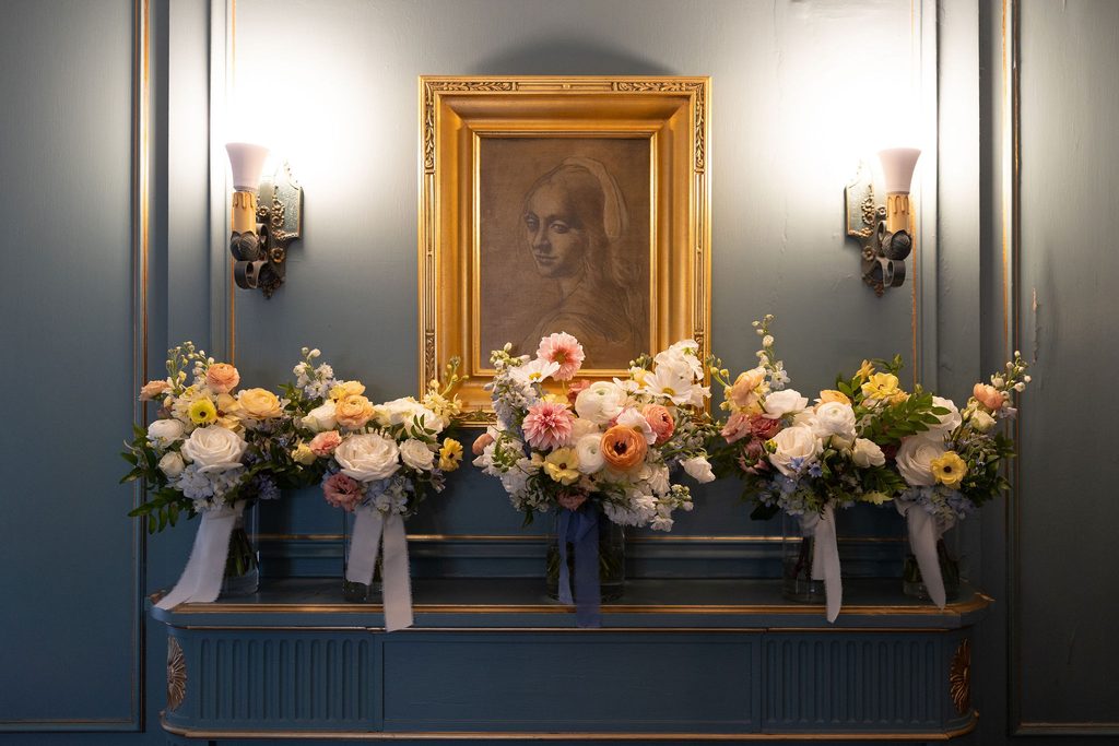 Spilling pastel floral arrangements displayed beneath a framed historic portrait inside an elegant room at Dresser Mansion.