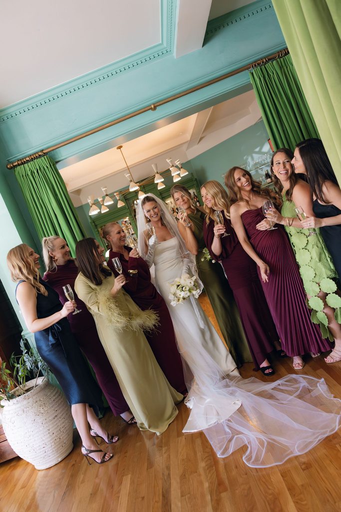 Bride surrounded by her bridesmaids in colorful dresses inside a brightly lit room at Bradford House with green curtains and framed artwork.