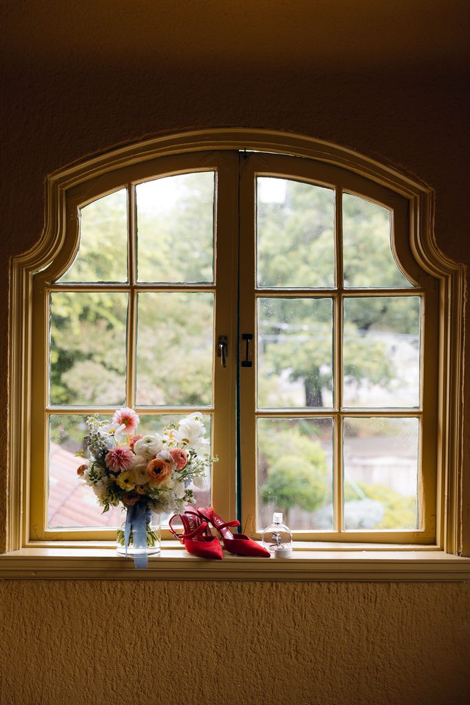Close-up of vibrant red bridal high heels placed on a stone window sill in front of a grand arched window at Dresser Mansion.