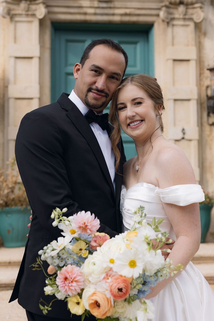 Bride and groom posing close together outdoors in front of a turquoise arched wooden doorway at Dresser Mansion in Tulsa.