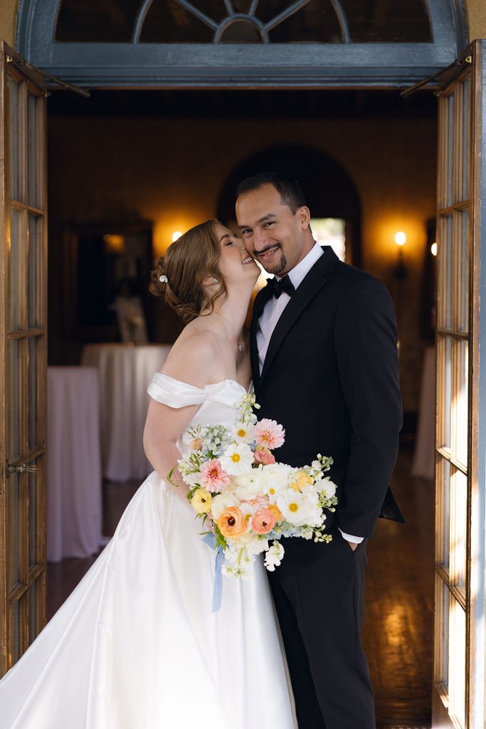 Bride and groom kissing indoors near warm wall sconces inside Dresser Mansion, with the bride holding a colorful pastel bouquet.