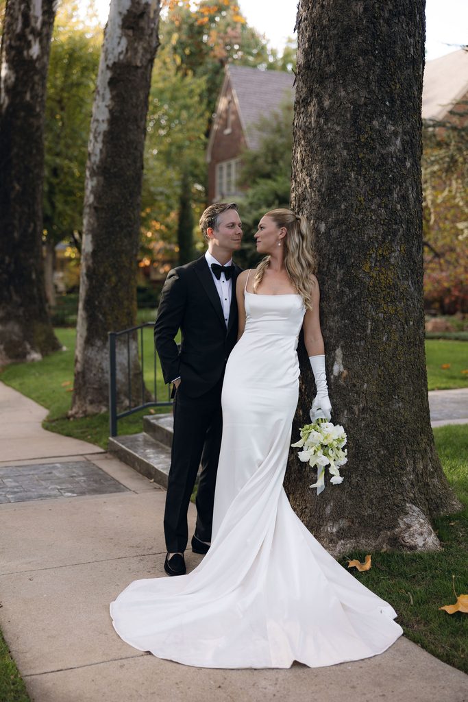 Bride in a strapless white gown with opera gloves holding a white bouquet, standing beside the groom in a black tuxedo on stone steps outside near trees.