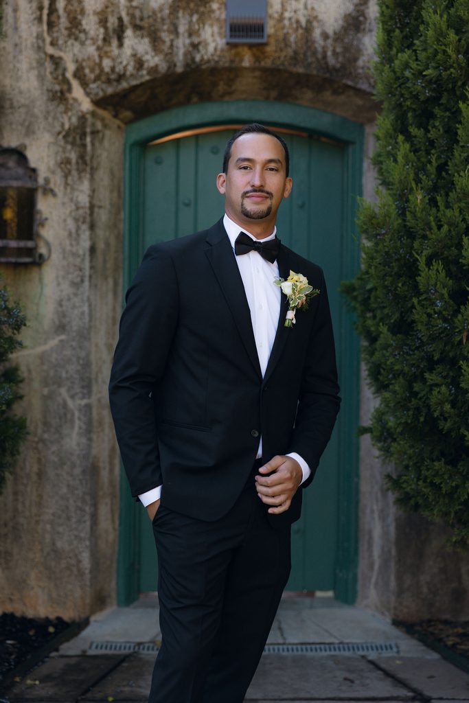 Groom wearing a classic black tuxedo with a white floral boutonniere, standing outside in front of a green arched wooden doorway at Dresser Mansion.