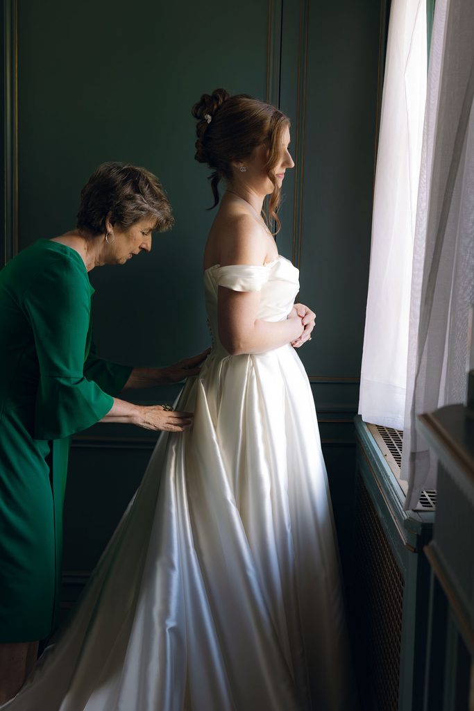 Bride standing by a window in an off-the-shoulder gown while an assistant in a emerald green dress helps adjust and zip up the back of her dress at Dresser Mansion.