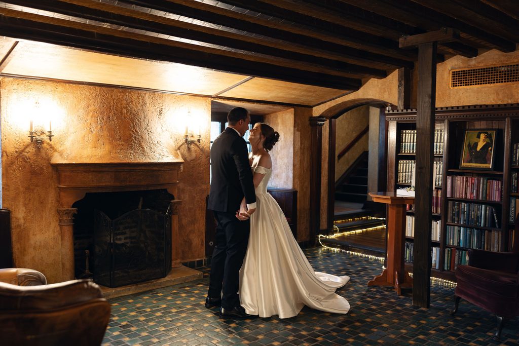 Bride and groom posing together inside an elegant room with glazed walls and a stone fireplace at Dresser Mansion in Tulsa.