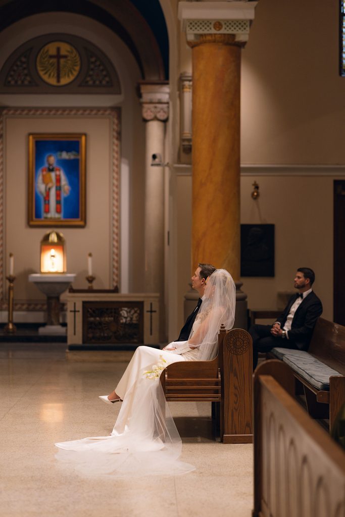 Bride in a white gown and long veil seated in a wooden pew inside the Cathedral of Our Lady of Perpetual Help, viewed from the aisle with arched sanctuary walls and a religious icon visible.