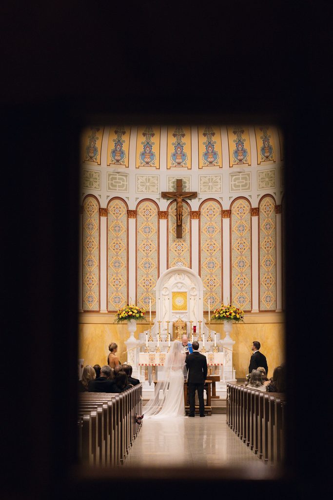 Bride in a white gown and long veil seated inside the Cathedral of Our Lady of Perpetual Help, viewed from the aisle with high arched sanctuary walls, tall columns, and a golden cross displayed on the altar.