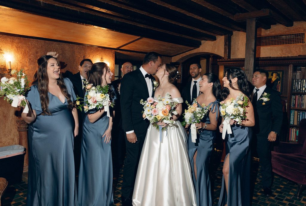 Bride and groom kissing outdoors surrounded by smiling bridesmaids in dusty blue dresses holding pastel bouquets on the grounds of Dresser Mansion.