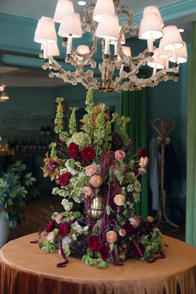 Lush, tall floral arrangement in rich burgundy, pink, and green hues displayed on a round table beneath a grand white chandelier inside Bradford House.