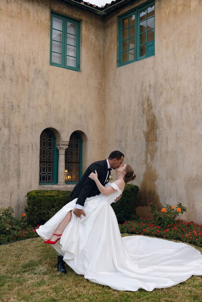 Groom dipping and kissing the bride wearing red heel shoes and a full ballgown on the lawn outside the stone exterior of Dresser Mansion in Tulsa, Oklahoma.