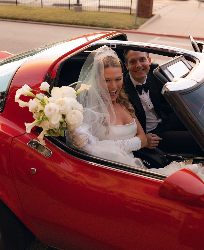 Bride in a white gown and long veil smiling alongside the groom inside a classic red Corvette getaway car with a white floral bouquet on the door.