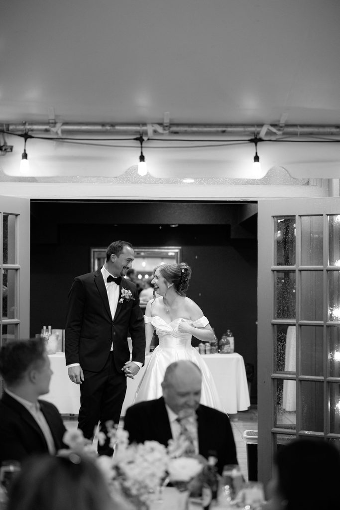 Black-and-white photo of the bride and groom smiling as they enter their reception room at Dresser Mansion, viewed from behind seated guests.