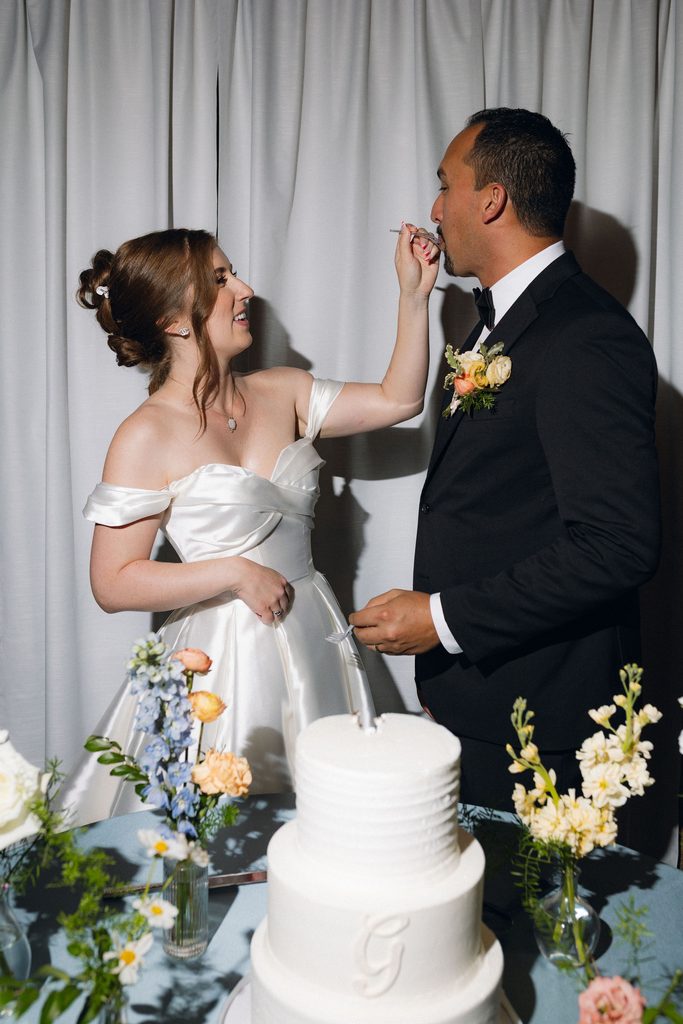 Bride in an off-the-shoulder wedding gown laughing as she feeds a piece of wedding cake to the groom next to a three-tiered white cake with pastel floral accents.