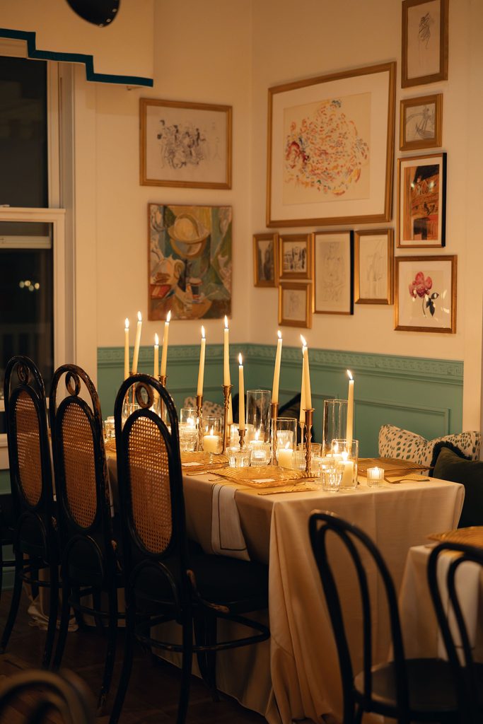 Warmly lit wedding reception dining setup inside a coral-pink room at Bradford House, featuring lit white taper candles along a wooden table with framed art hanging on the walls.